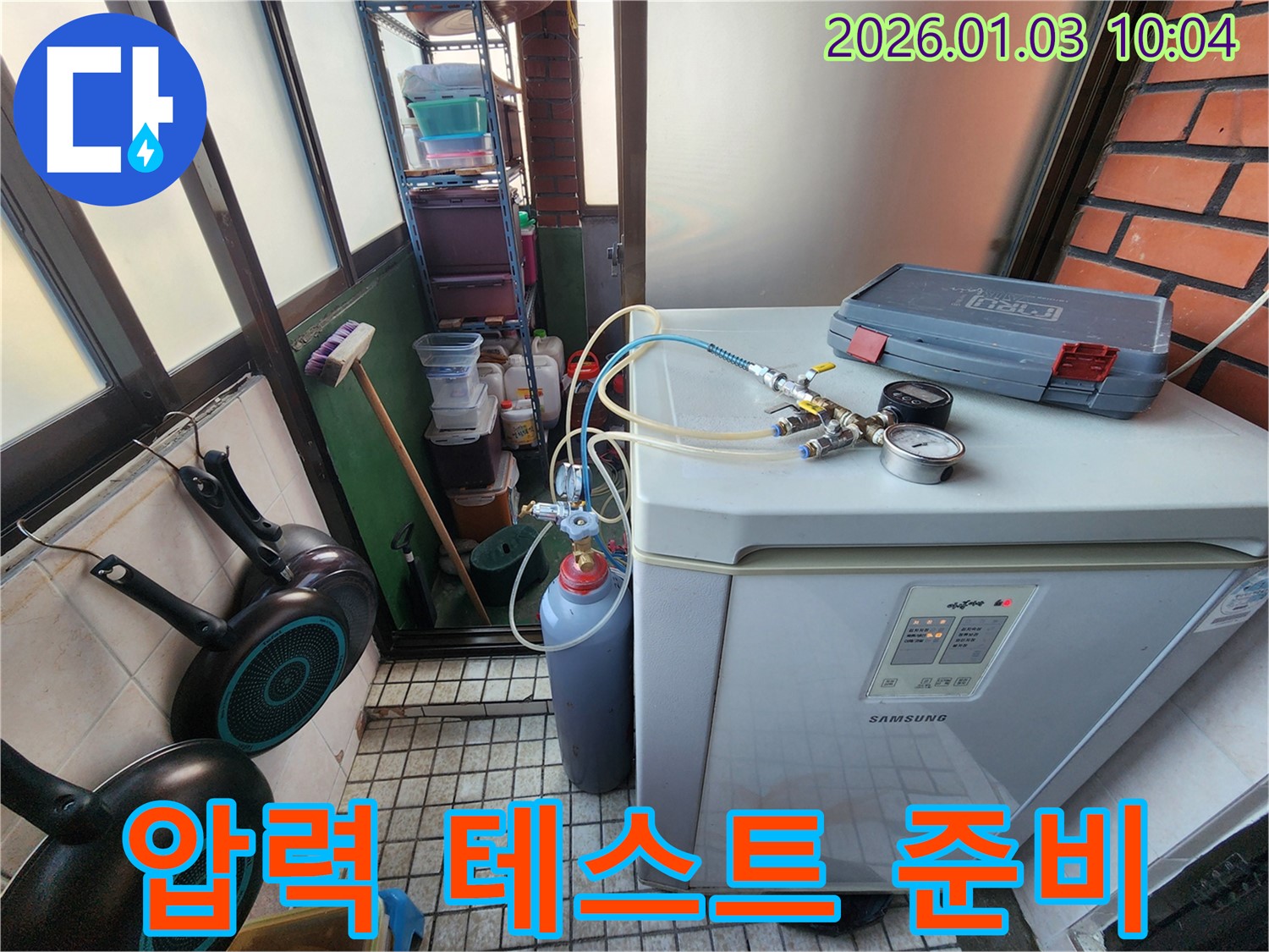 누수 탐지를 위해 냉장고 옆 공간에서 압력 테스트 장비를 설치하고 있습니다 - 꼼꼼한 준비가 정확한 진단의 시작입니다.