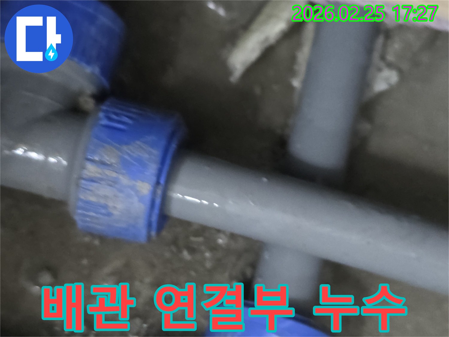 이 이미지는 주택 내부의 수도 배관 연결 부위에서-물이 새는 현장을 보여주며-회색 파이프와 파란색 부품이 특징입니다.
