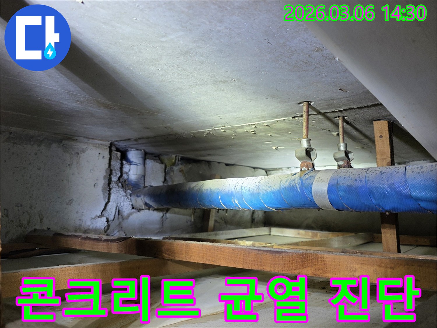 지하 공간의 파란색 배관 옆-심각하게 균열된 콘크리트 벽면을-누수 엔지니어가-구조적 안정성 진단을 위해-점검하는 모습