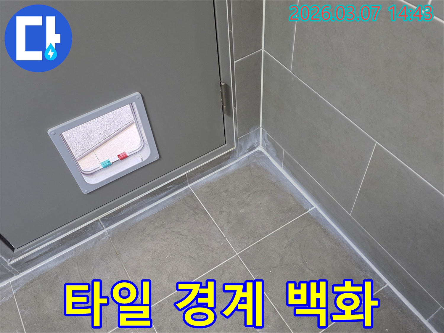 회색 타일 바닥과 벽면의 이음새-그리고 문 주변에 나타난-흰색 백화 현상은-내부로 물이 스며들고 있음을 시사하는 누수 증상입니다.