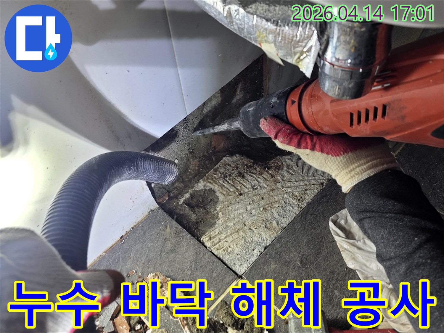 누수 탐지 후 바닥 타일을 해체하는 작업자가 드릴을 사용하며-파손된 타일 조각과 흡입 호스가 보이는 공사 현장