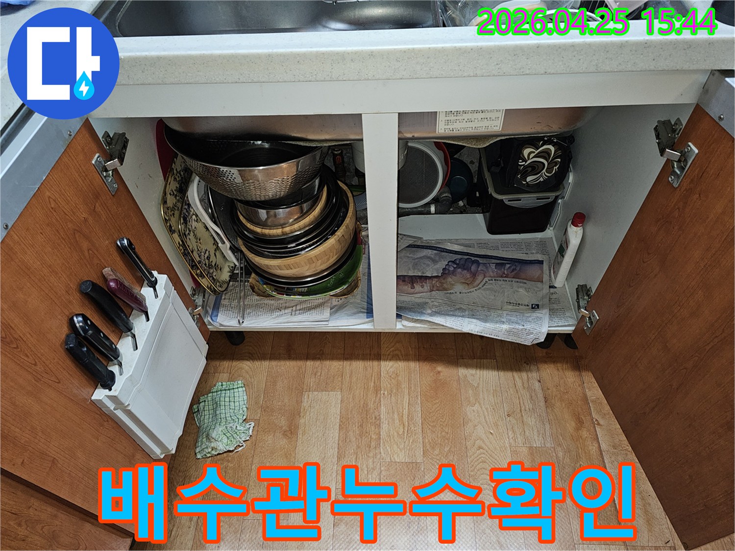 싱크대 하부장 내부의 모습-누수 점검을 위해-그릇과 배관이 보이고 바닥에 신문지가 깔려 있습니다.