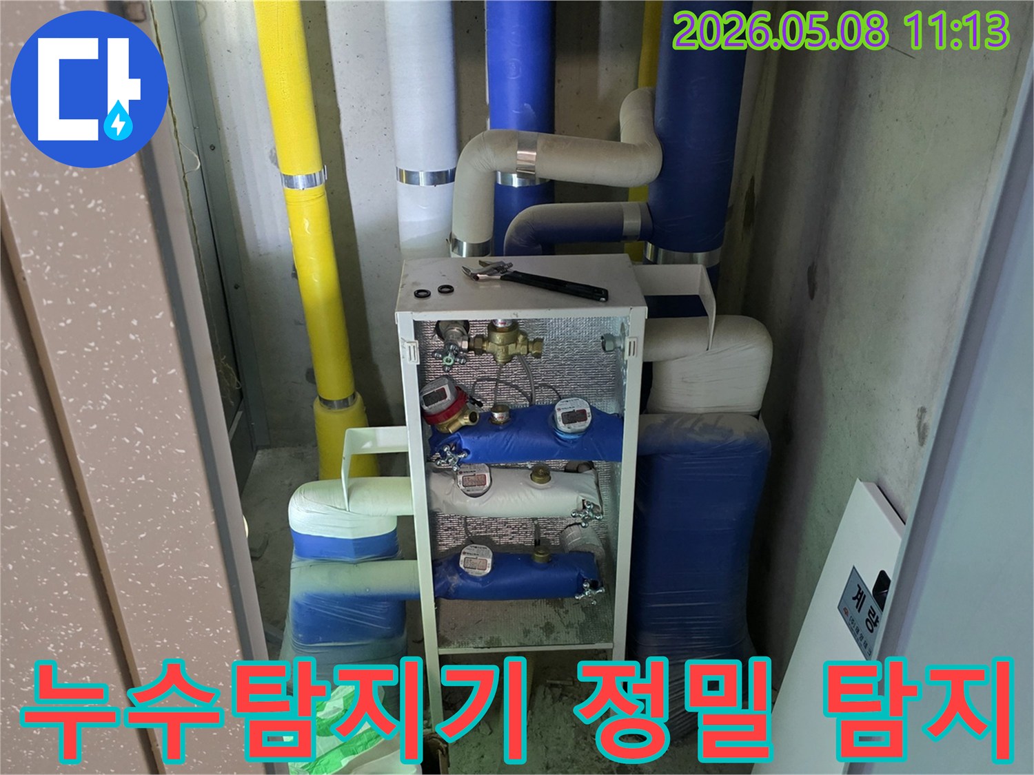 이 이미지는 건물의 유틸리티 공간에 설치된-다양한 배관과 계량기-그리고 작업 도구를 보여주며-이는 누수탐지기를 활용한-정밀 탐지 작업이 진행될 준비를 나타냅니다