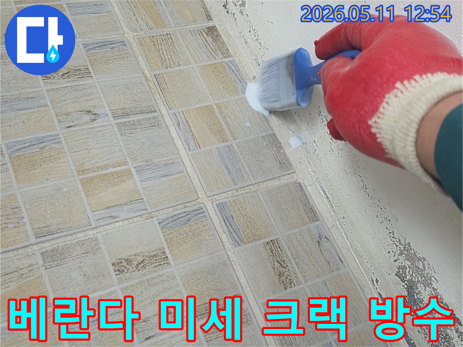 붉은 장갑을 낀 작업자가-베란다 벽면 하단의 미세 크랙과 타일 경계에-방수액을 붓으로 꼼꼼히 바르며-누수를 차단하고 있습니다