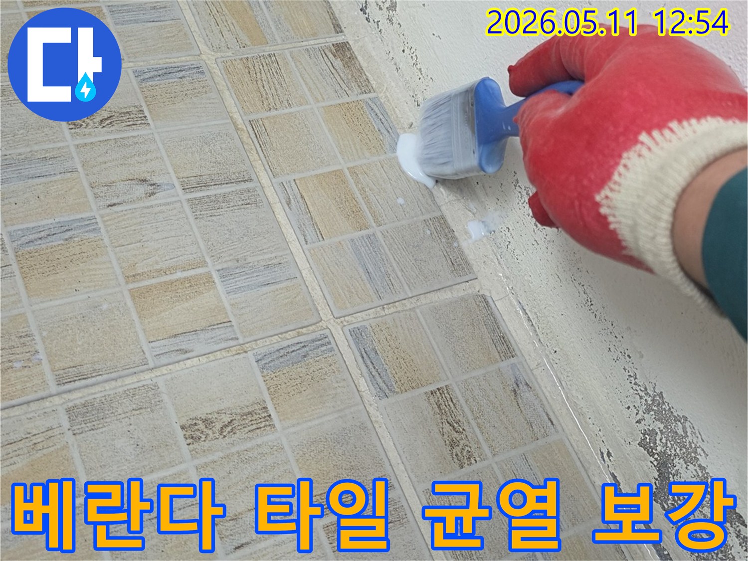 붉은 장갑을 낀 작업자가-베란다 타일의 균열 부위와 벽면 경계에-방수액을 붓으로 꼼꼼히 바르며-누수를 방지하고 있습니다