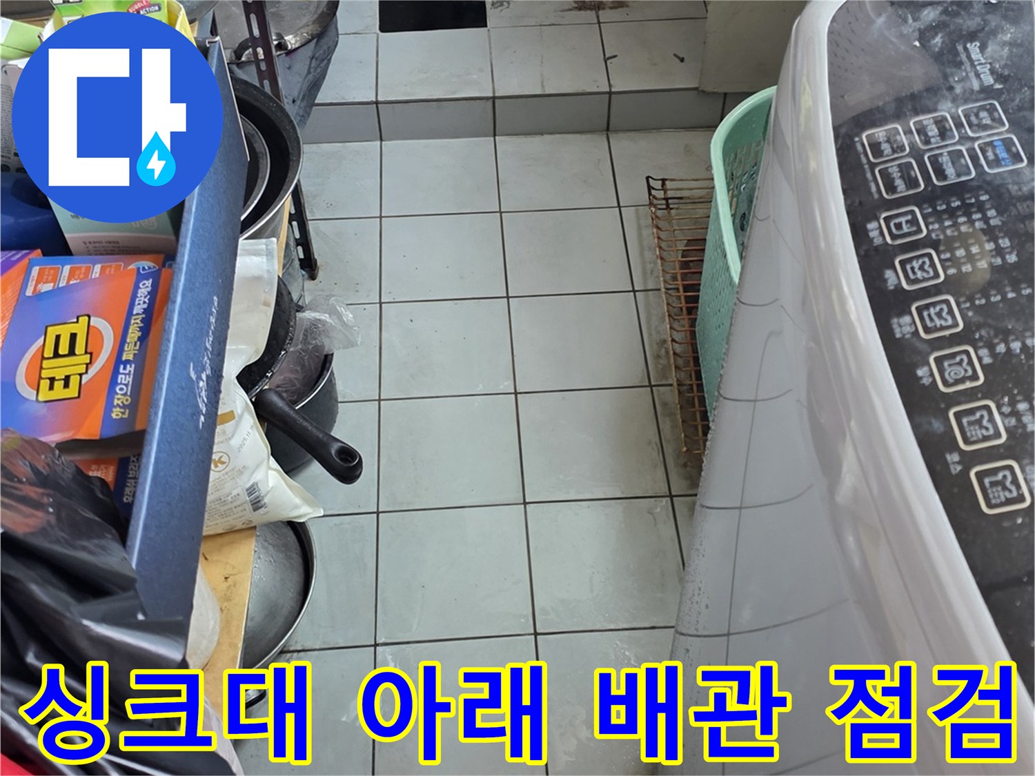 주방 싱크대 하단부의 복잡한 배관과 주변 물품들을 찍은 이미지입니다.