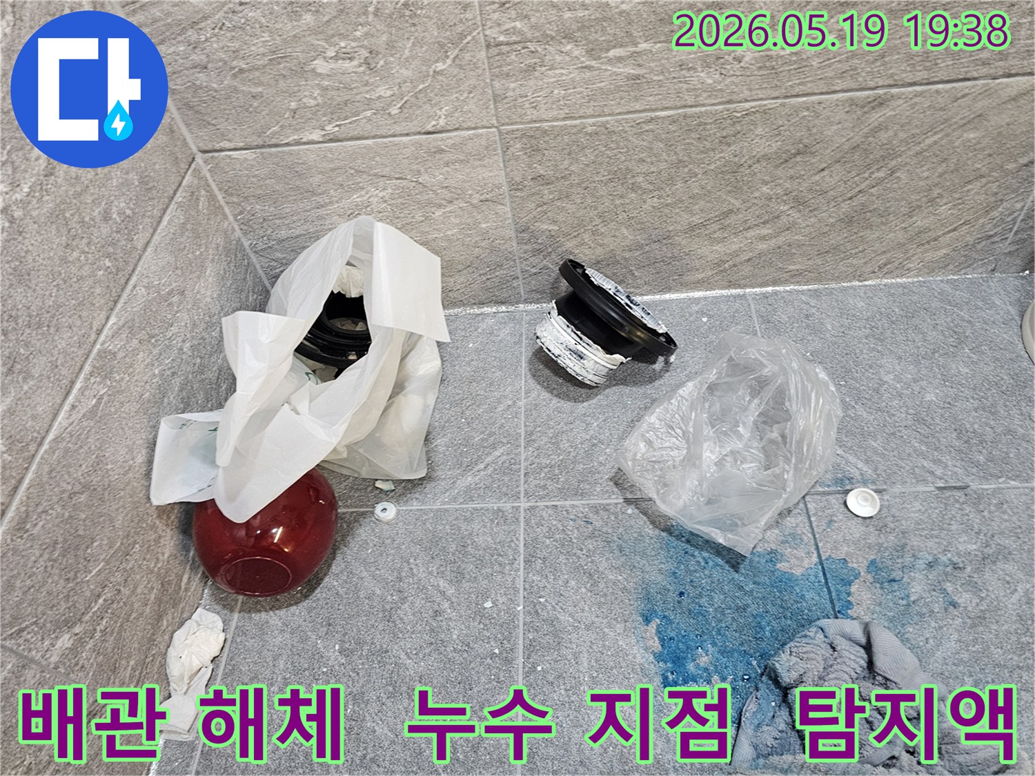누수 공사 현장에서-파란색 탐지액으로 확인된 누수 지점과-교체를 위해 해체된 배관 부품들이 바닥에 놓여 있습니다