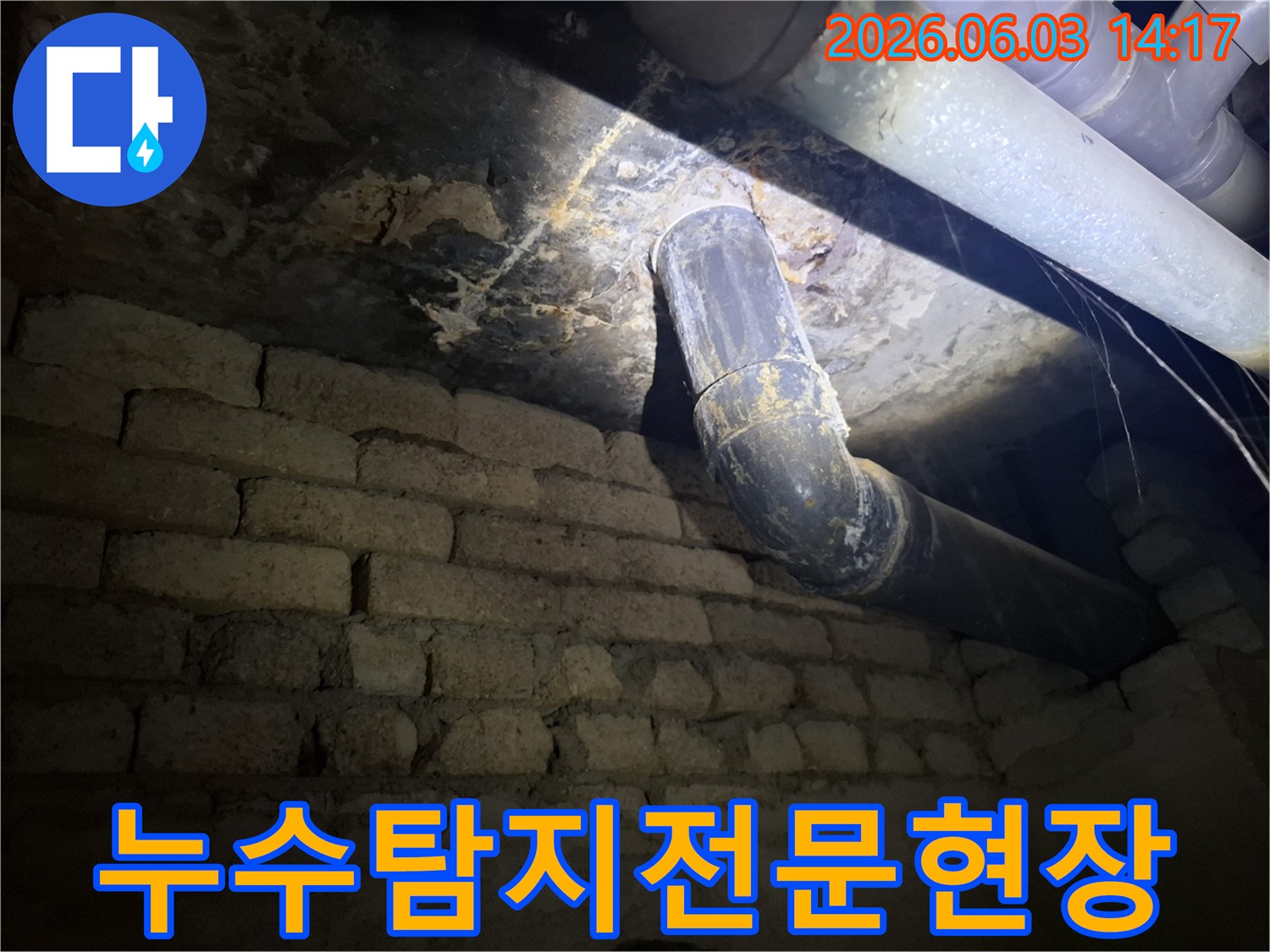 지하의 낡은 배관에서-물이 새어 나와-벽돌 벽에 스며든-누수 자국을-조명으로 비추어-면밀히 검사하는 모습