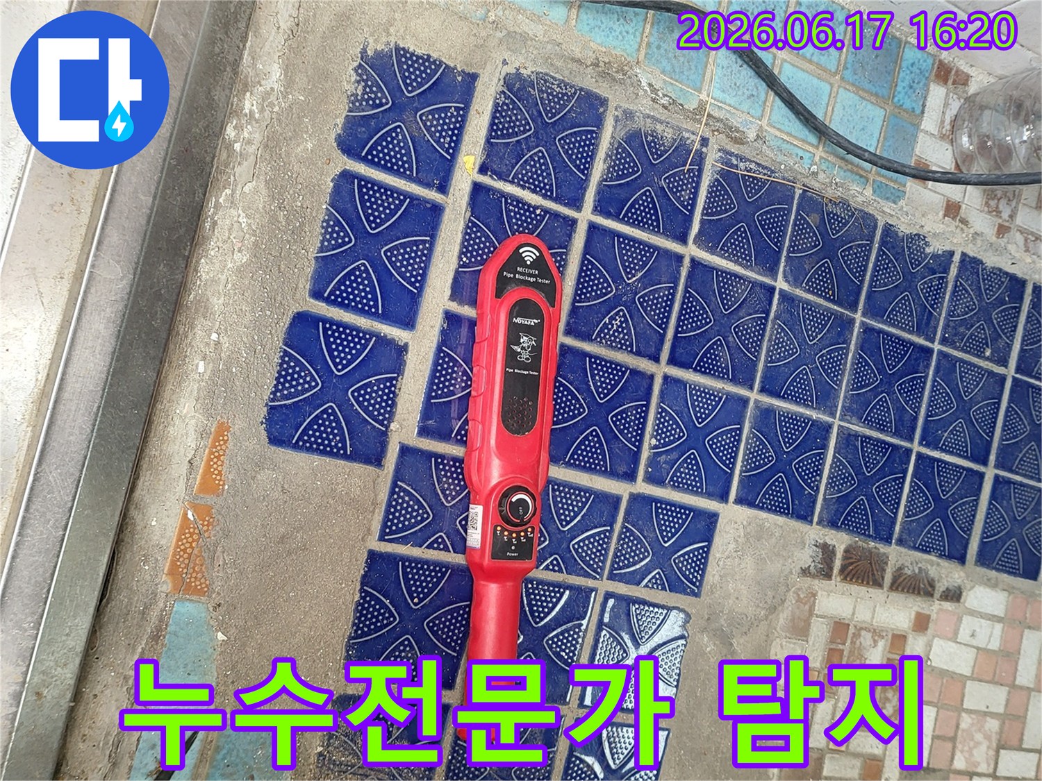 파란색 타일이 뜯겨나간 욕실 바닥에 놓인-붉은색 누수탐지 장비가-전문가의 손길로 누수 지점을 찾아내는 모습