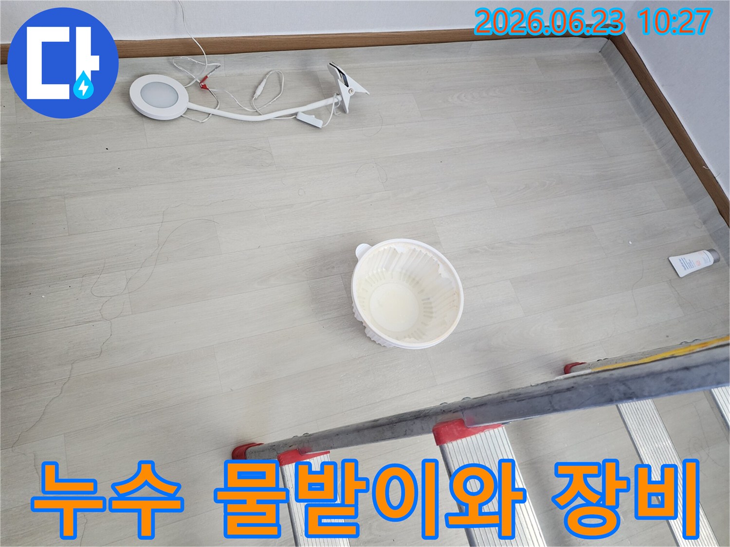 천장에서 물이 떨어져 바닥에 놓인 물받이 그릇과-누수 지점 확인 및 작업을 위한 사다리-조명 장비가 함께 보이는-누수 현장 준비 단계의 모습입니다