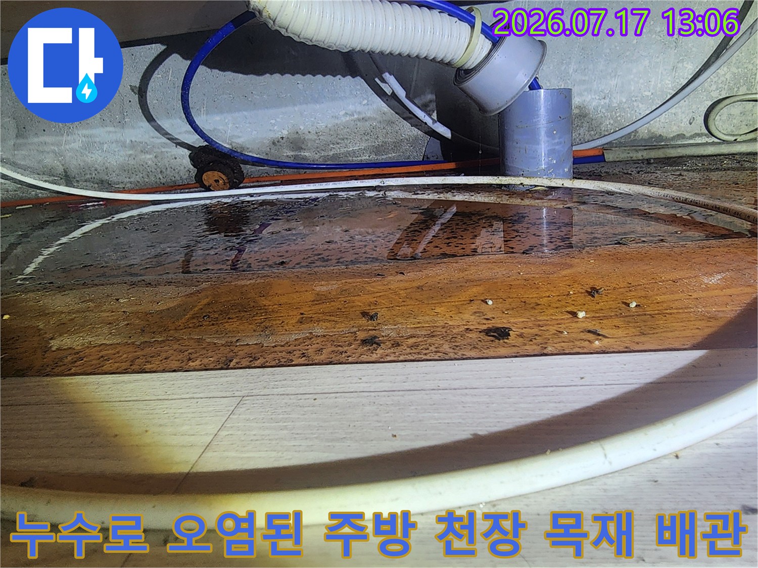 물에 젖어 갈색으로 변한 주방 천장의 나무 판넬과 그 아래 배치된 흰색 및 파란색 배관들