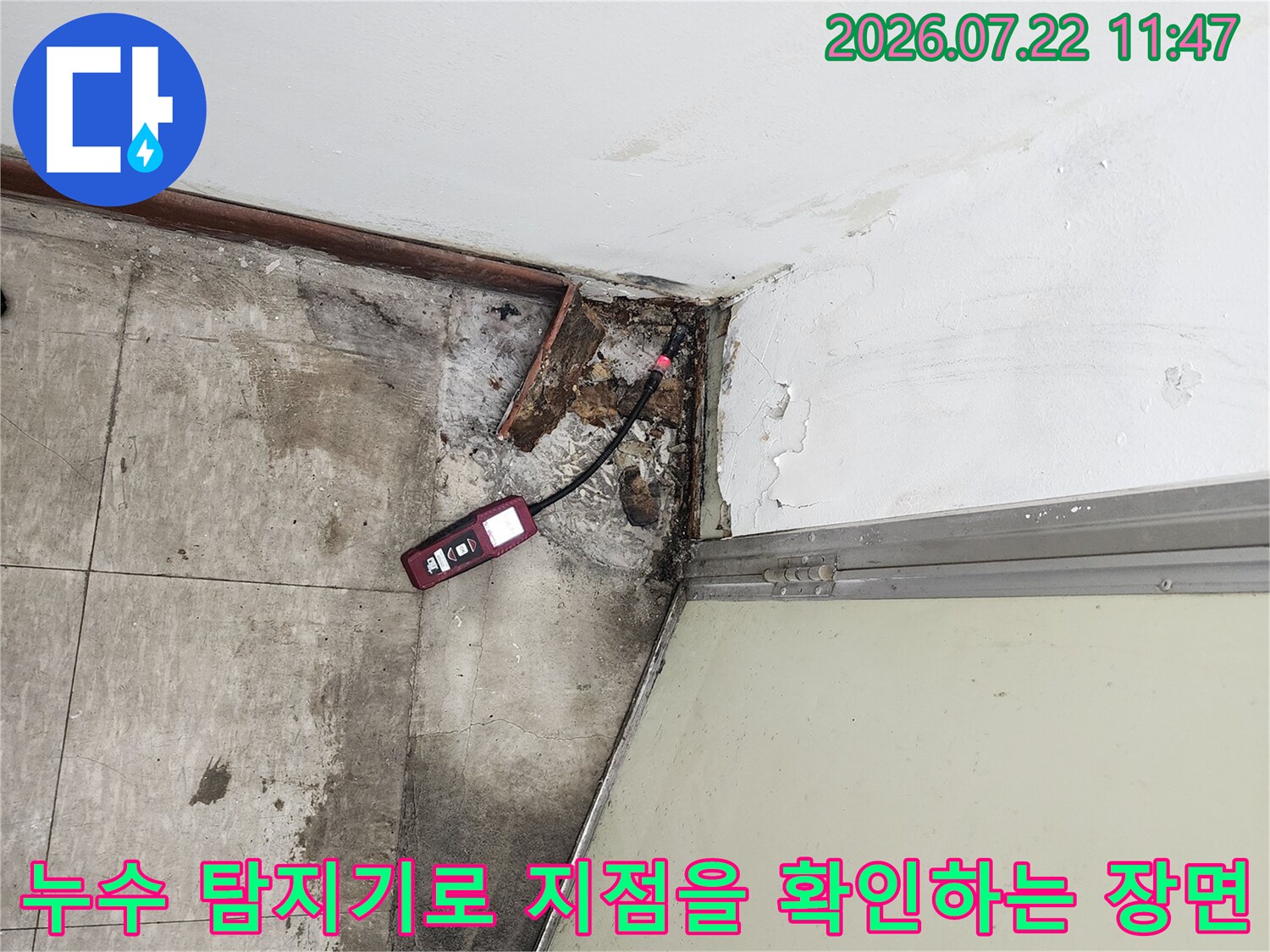 타일 바닥의 젖은 얼룩과 벽면 하단 파손 부위-빨간색 누수 탐지 장비가 놓여 있음