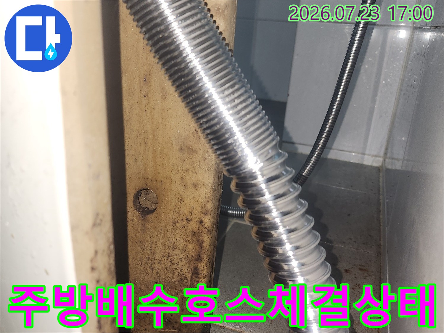 싱크대 하부장의 은색 금속 주름 호스가 벽면 배수구에 꽂혀 있는 장면