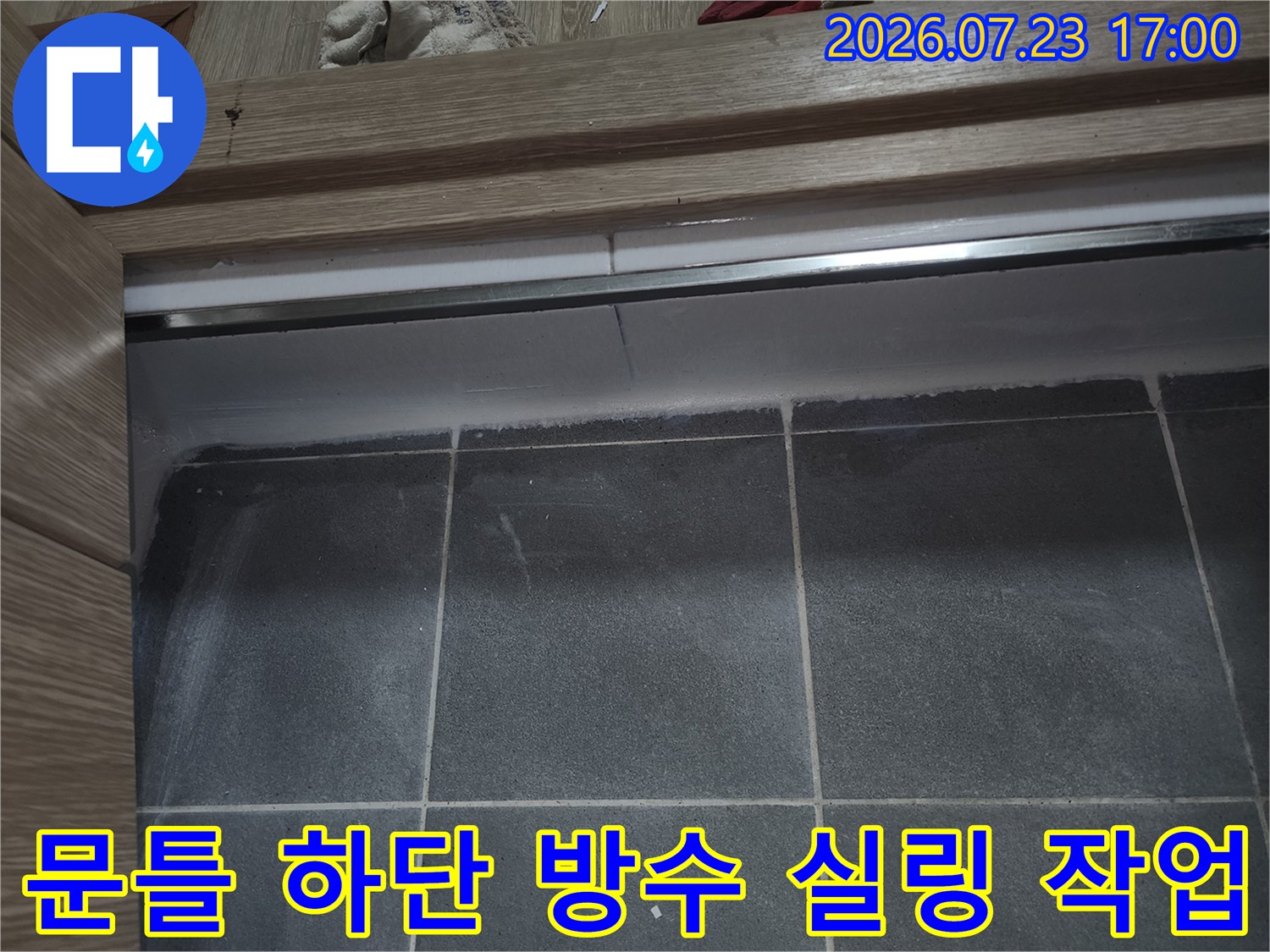 회색 타일 바닥과 목재 문틀 경계면에 도포된 백색 실리콘 마감재