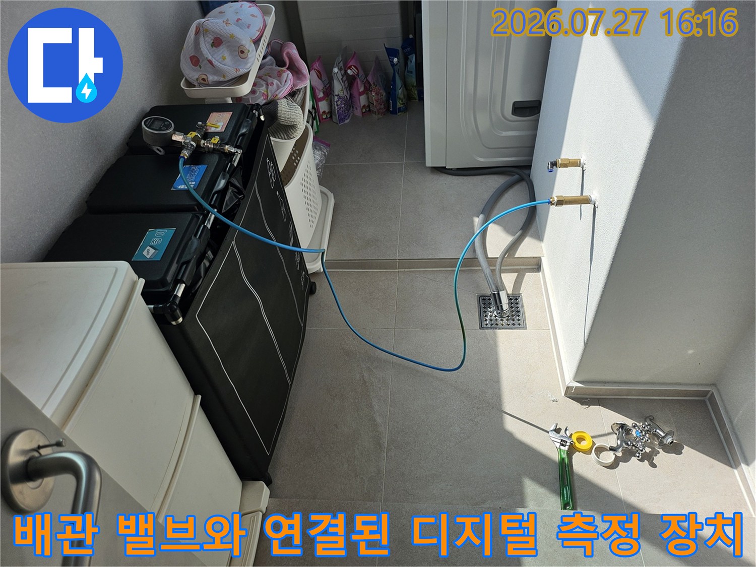 디지털 압력계가 부착된 검은색 장비와 파란색 호스가 벽면 배관 밸브에 연결되어 점검 중인 사진