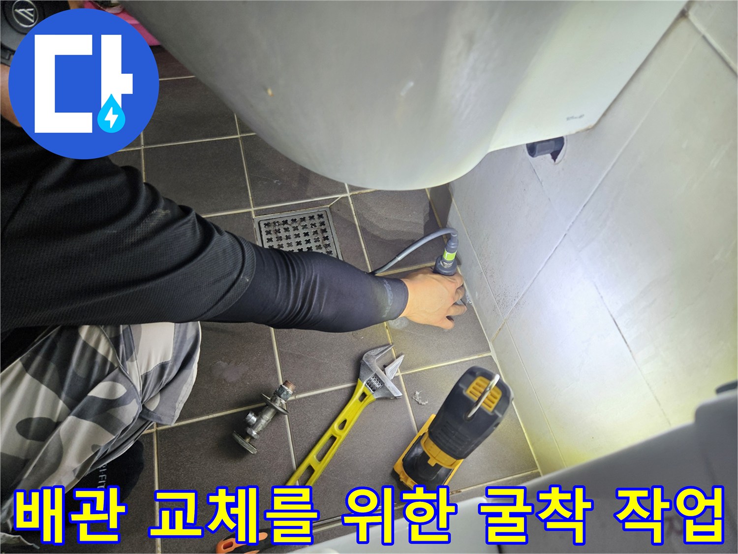 전동 드릴과 공구들을 활용하여 화장실 벽면 내부 배관을 확인하기 위해 타일을 제거하는 장면입니다.